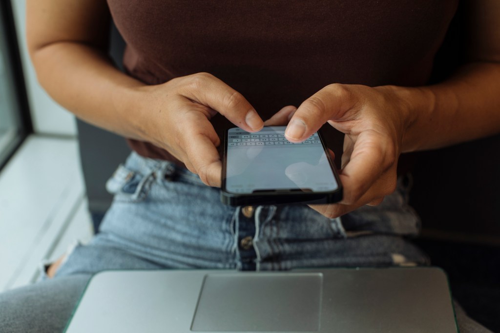 Woman using a smartphone while holding a laptop on her lap