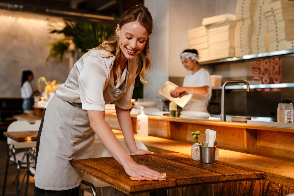 Young white woman wearing apron cleaning table while working in restaurant.