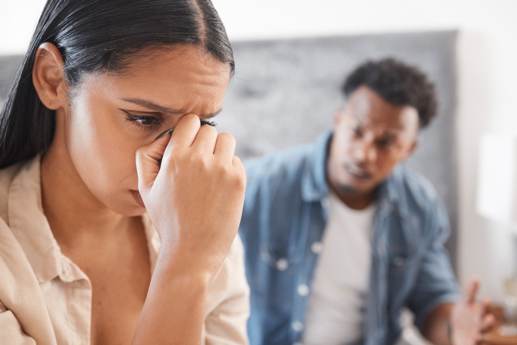 A couple is fighting fiercely on a bed while a woman covers her face in distress. Interracial couples fighting and divorce issues portrayed.