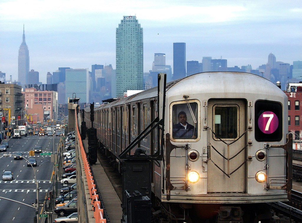 Front view of a 7 train as it moves on elevated tracks with the Manhattan skyline behind it. 