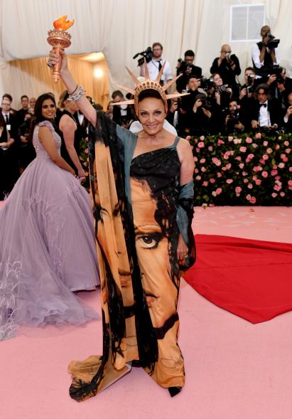 Diane von Furstenberg at Costume Institute benefit gala in blue/green dress with gold bangle and earrings. Wearing metallic statue on head.