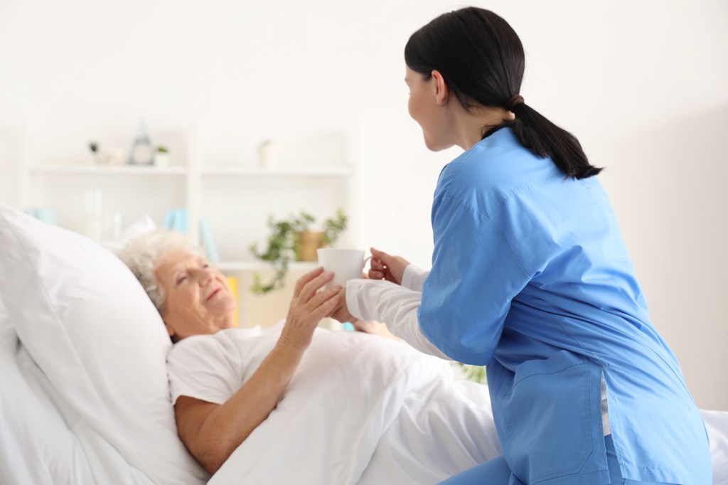 A nurse giving a cup of tea to a senior woman in a bedroom