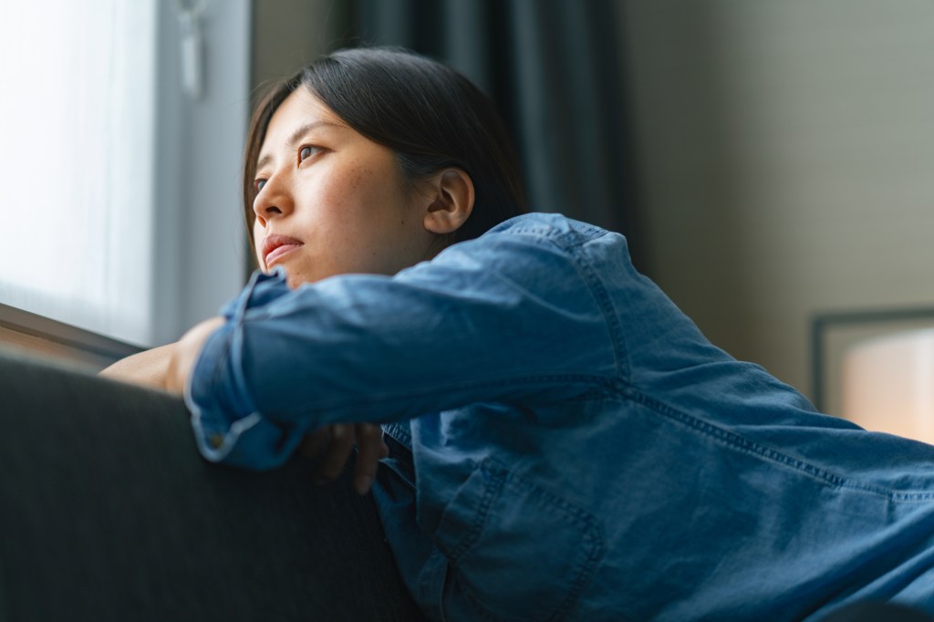 A young woman is sitting on a sofa by the window and looking outside through the window at home.