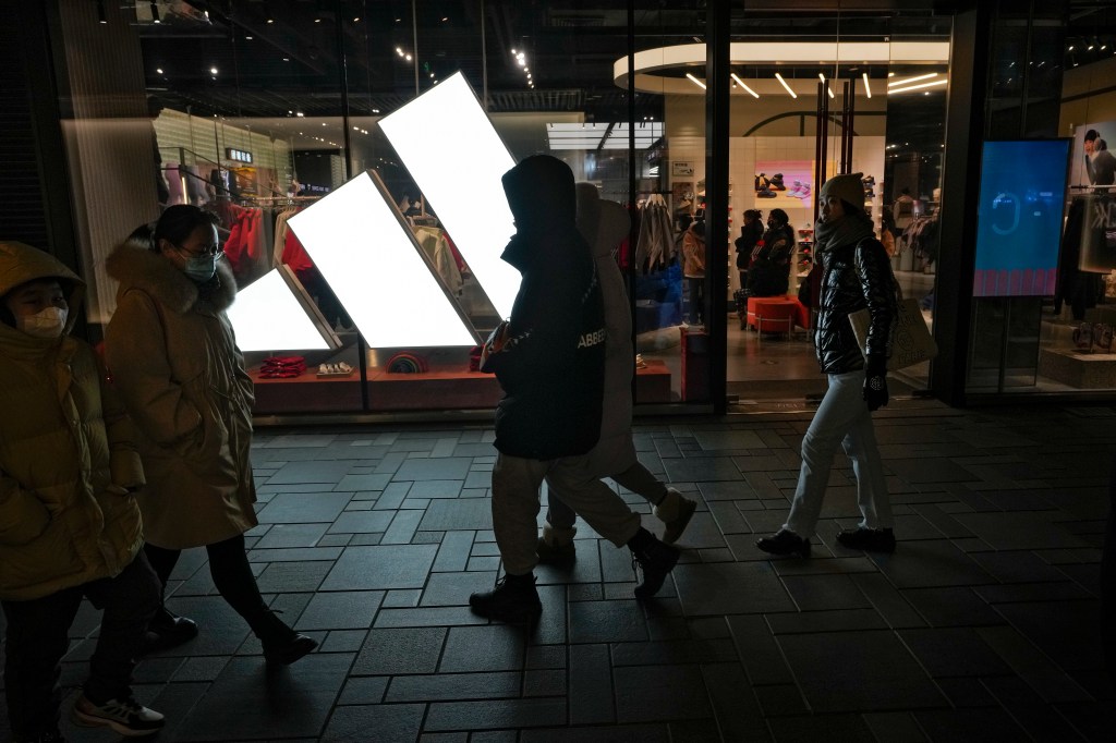 People walking in front of an Adidas store at an outdoor mall in Beijing, with a clear blue sky in the background. No celebrity faces detected.