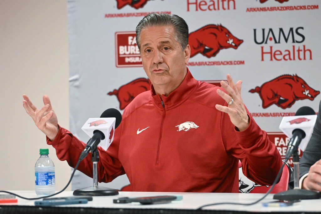 John Calipari in a red jacket, answering questions from reporters after being announced as the new Arkansas men's basketball coach in 2024