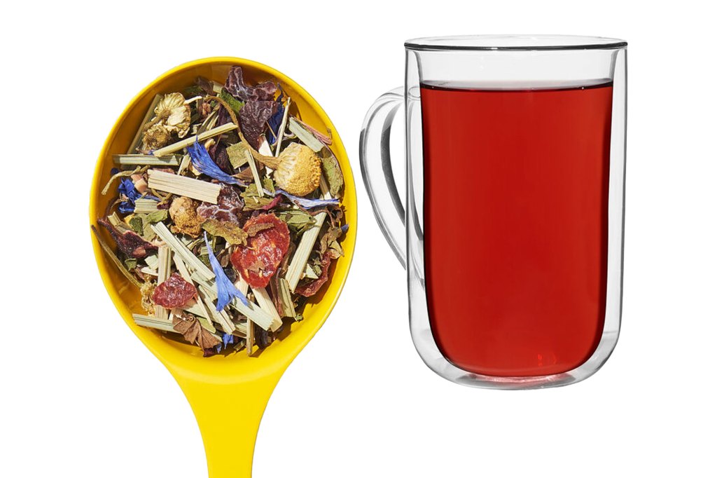 A spoon with dried herbs next to a glass of red liquid