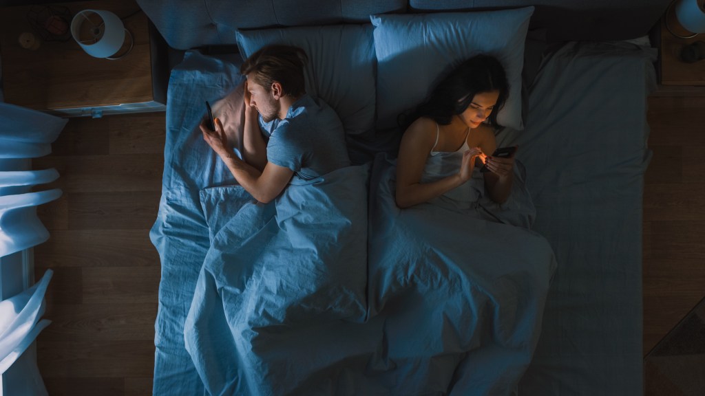 Millennial couple in bed, turned away from each other, engrossed in their smartphones