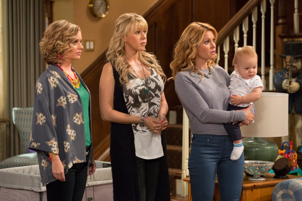 A group of women, Andrea Barber, Jodie Sweetin, Candace Cameron Bure, standing in a room with a baby from the TV show 'Fuller House'.