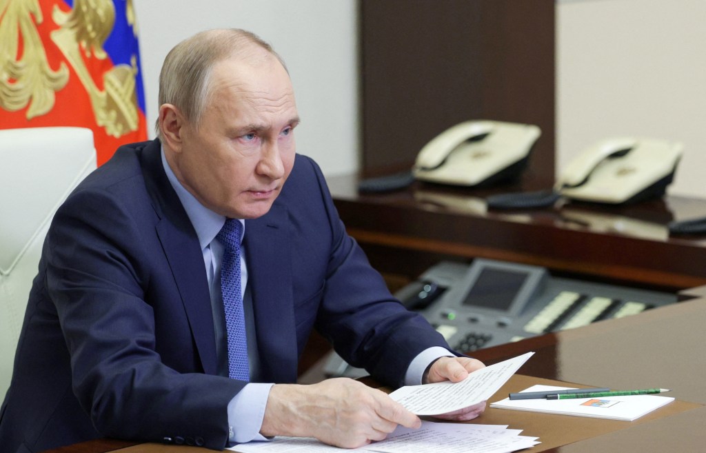 Vladimir Putin at a desk, wearing a blue suit with blue tie, holding papers