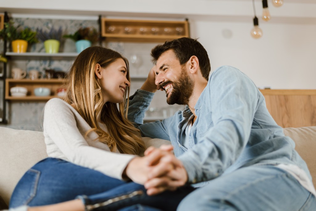 A loving happy couple sitting on a couch at home, holding hands and talking to each other