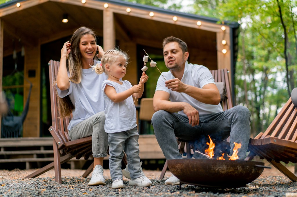 A family roasting marshmallows by a fire pit in the forest near their country house