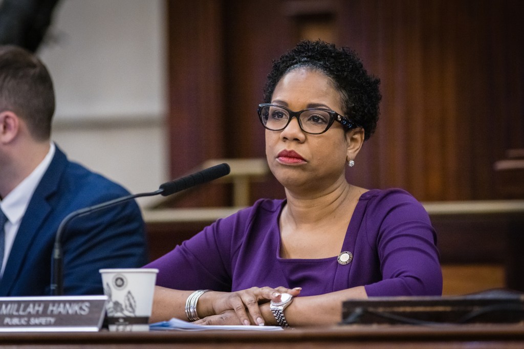 Councilwoman Kamillah Hanks in a purple top and black rimmed glasses sitting at a microphone at the city council