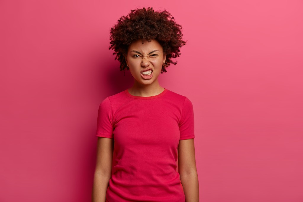 Annoyed woman in casual t-shirt clenching teeth and looking displeased at the camera against a pink background.