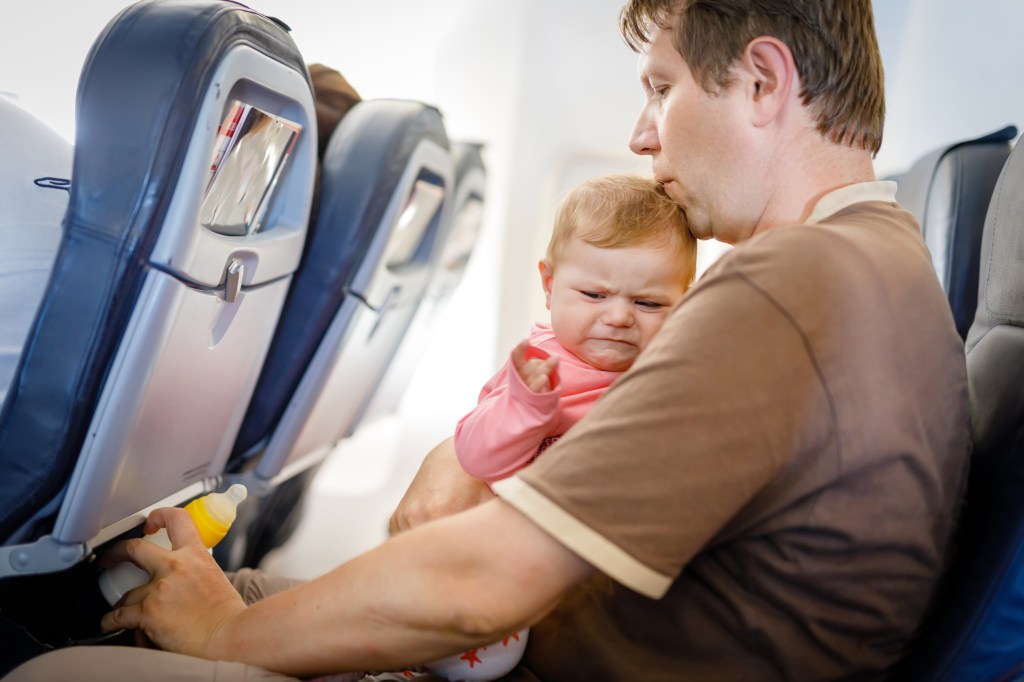 Young tired father and his crying baby daughter during flight on airplane going on vacations. Dad holding baby girl on arm. Air travel with baby, child and family concept.