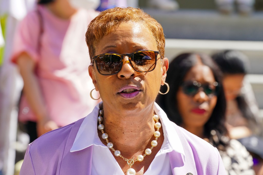 Adrienne Adams in a purple top and blazer with gold hoop earrings, a large pearl and gold necklace, speaking and looking into the camera while wearing brown sunglasses outside