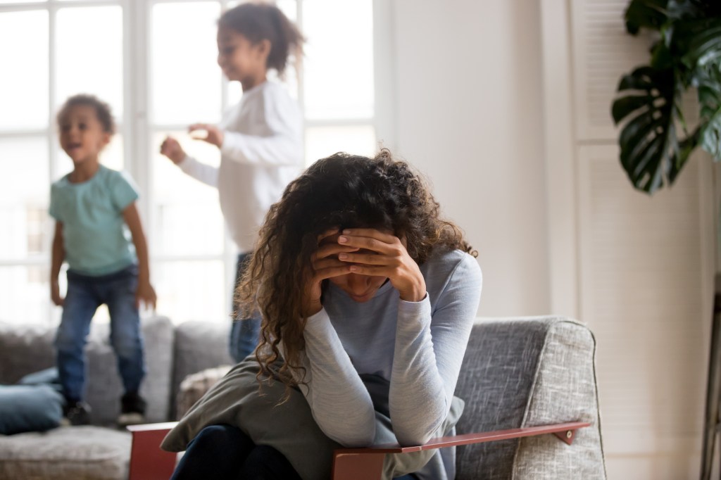 Black single frustrated woman hold her head with hands sitting on chair in living room, playful kids jumping on couch on a background. Tiredness, depression difficult to educate children alone concept