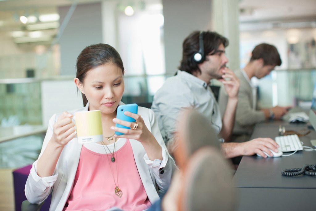 Businesswoman Crystal Yu relaxing in office, texting on her phone and drinking coffee