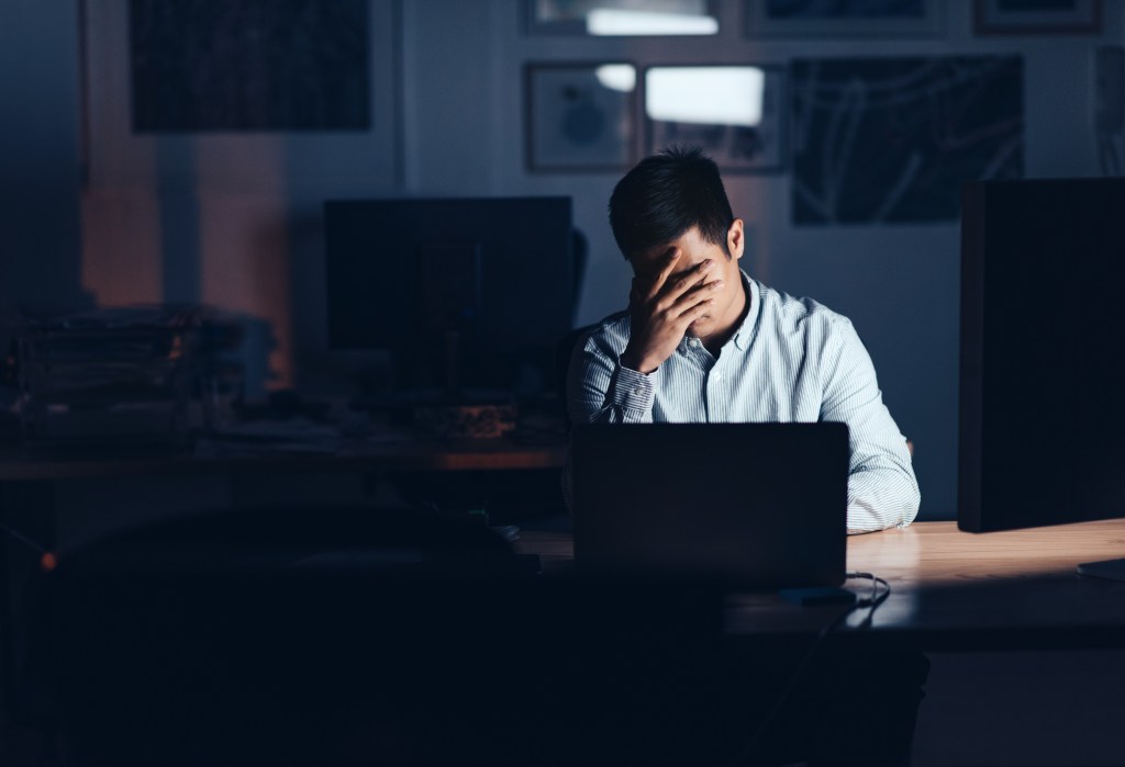 Young Asian businessman looking exhausted with his head in his hand while working alone at his desk in an office late at night