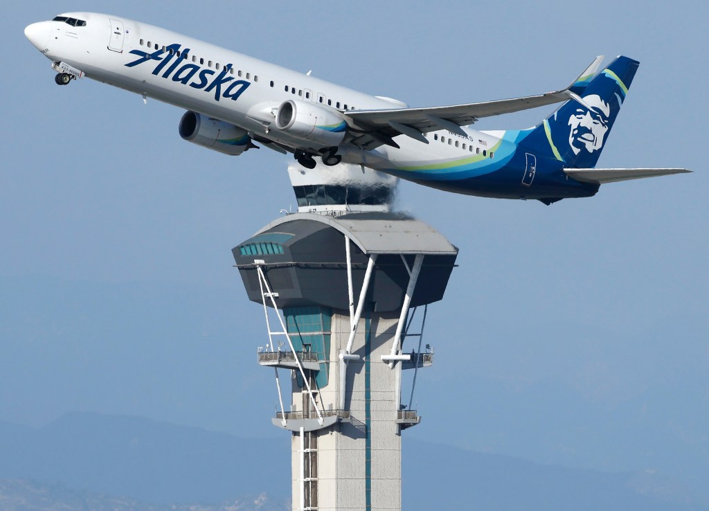 An Alaska Airlines plane takes off over an air traffic control tower.