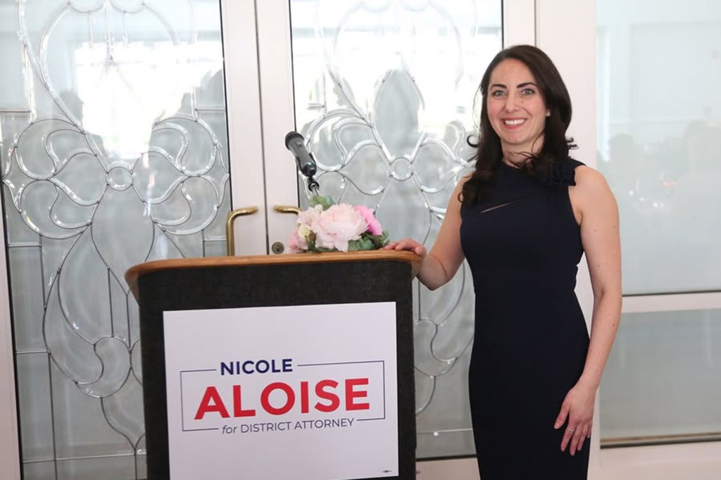 Nicole Aloise smiling, standing next to a podium with her campaign sign, "Nicole Aloise for District Attorney."