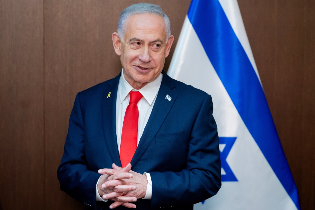 Benjamin Netanyahu speaks while wearing a blue suit, red tie, and yellow ribbon, with an Israeli flag in the background.