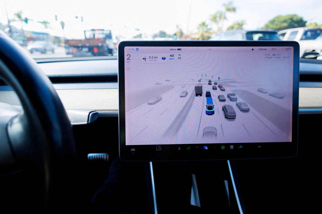 A Tesla Model 3 car's dashboard screen displays its self-driving interface, showing a blue car on a digital highway with other cars, next to a partially visible steering wheel.
