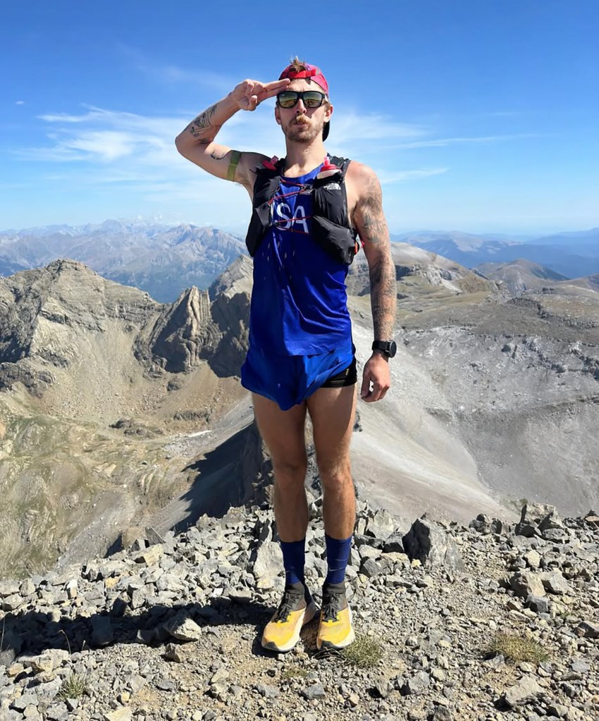 Man saluting from a mountain top, wearing a blue USA running top and running vest.