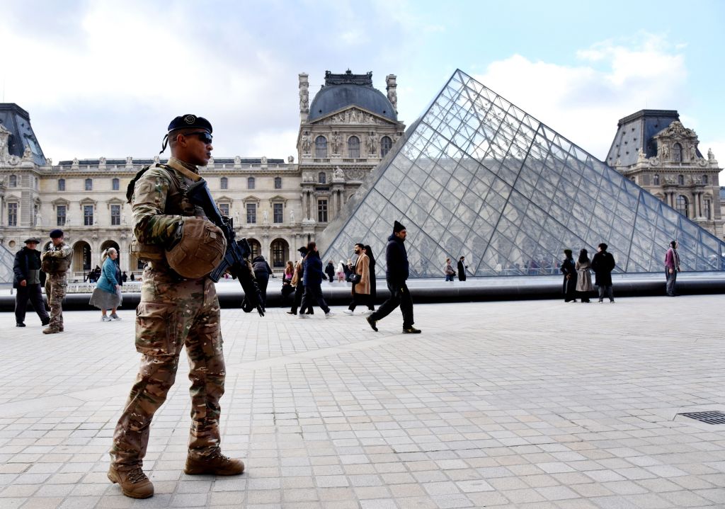 A soldier in camouflage uniform and sunglasses stands guard near the Louvre Museum, with the museum building and glass pyramid in the background.