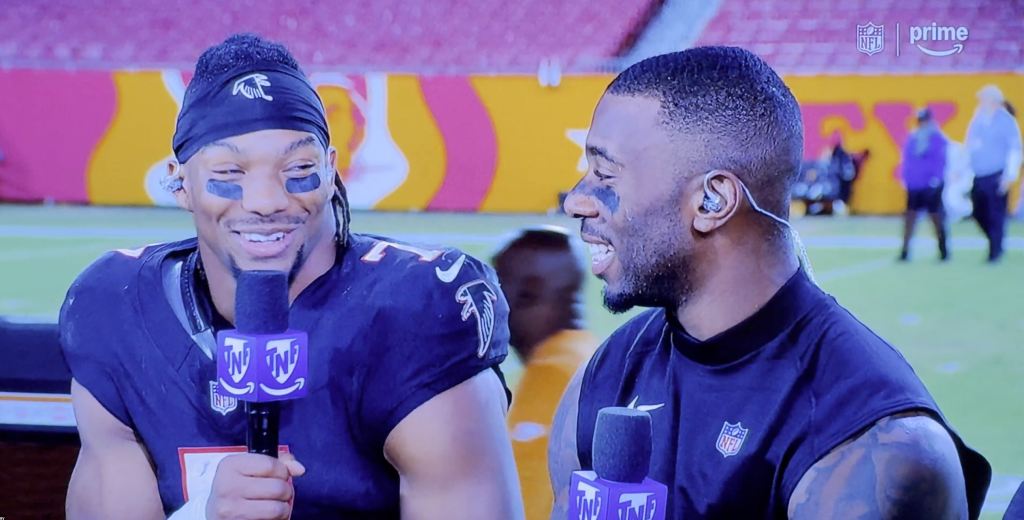 Two football players in dark jerseys smiling, with one holding a microphone with the NFL and Prime logos.