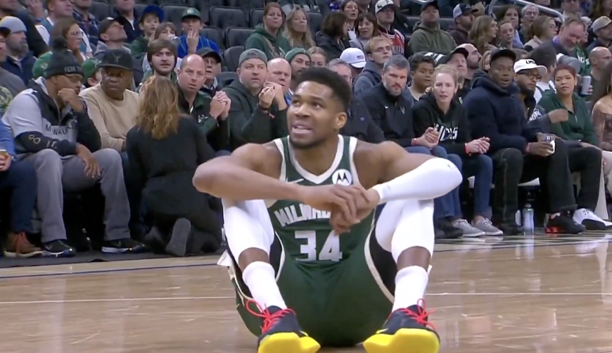 Basketball player Giannis Antetokounmpo sitting on the court during a game.