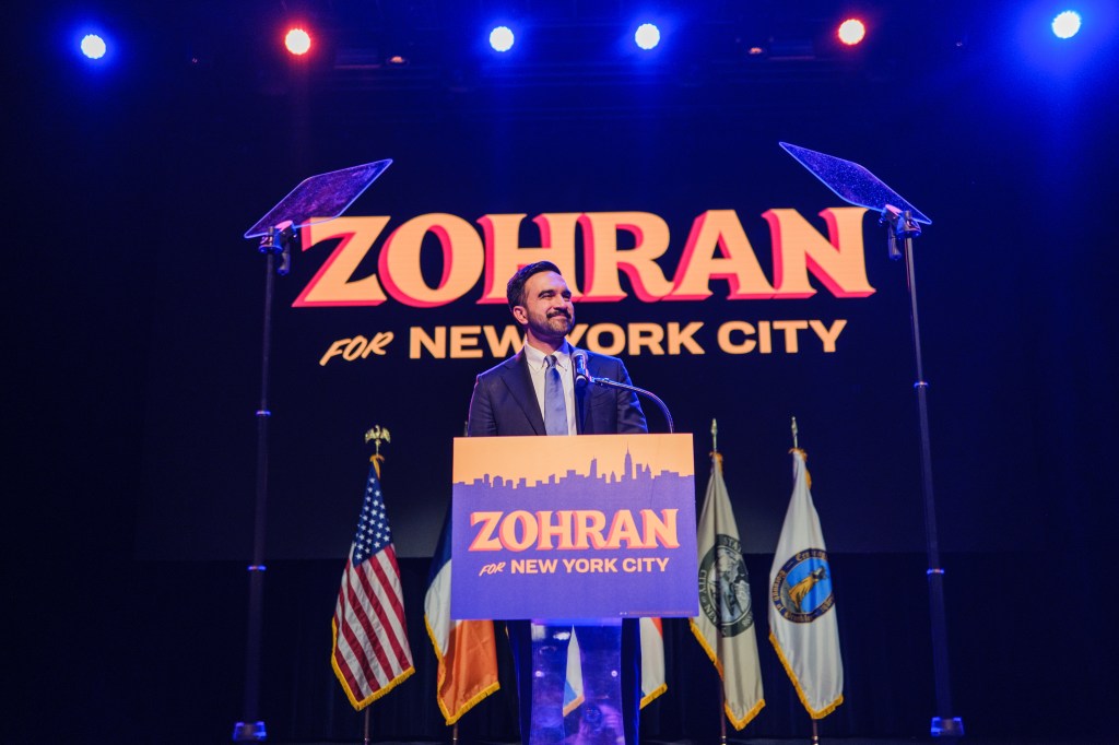 Zohran Mamdani celebrates after securing his win for mayor of New York City, speaking at a podium with “Zohran for New York City” on it and an American flag to his left.
