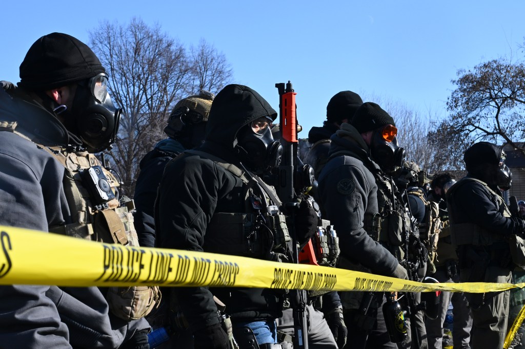 Federal agents in gas masks and tactical gear behind yellow "Police Line Do Not Cross" tape.