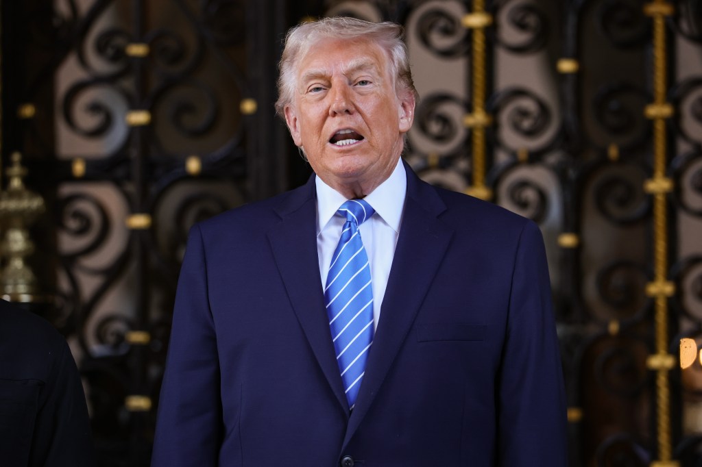 Donald Trump speaking with his mouth open, wearing a blue suit and striped blue tie, in front of a dark, ornate gate.