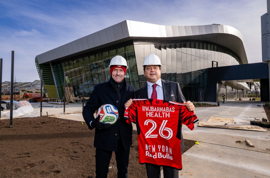 Two men in hard hats pose outside the Red Bull New York Training Facility, one holding a soccer ball and the other a New York Red Bulls jersey.