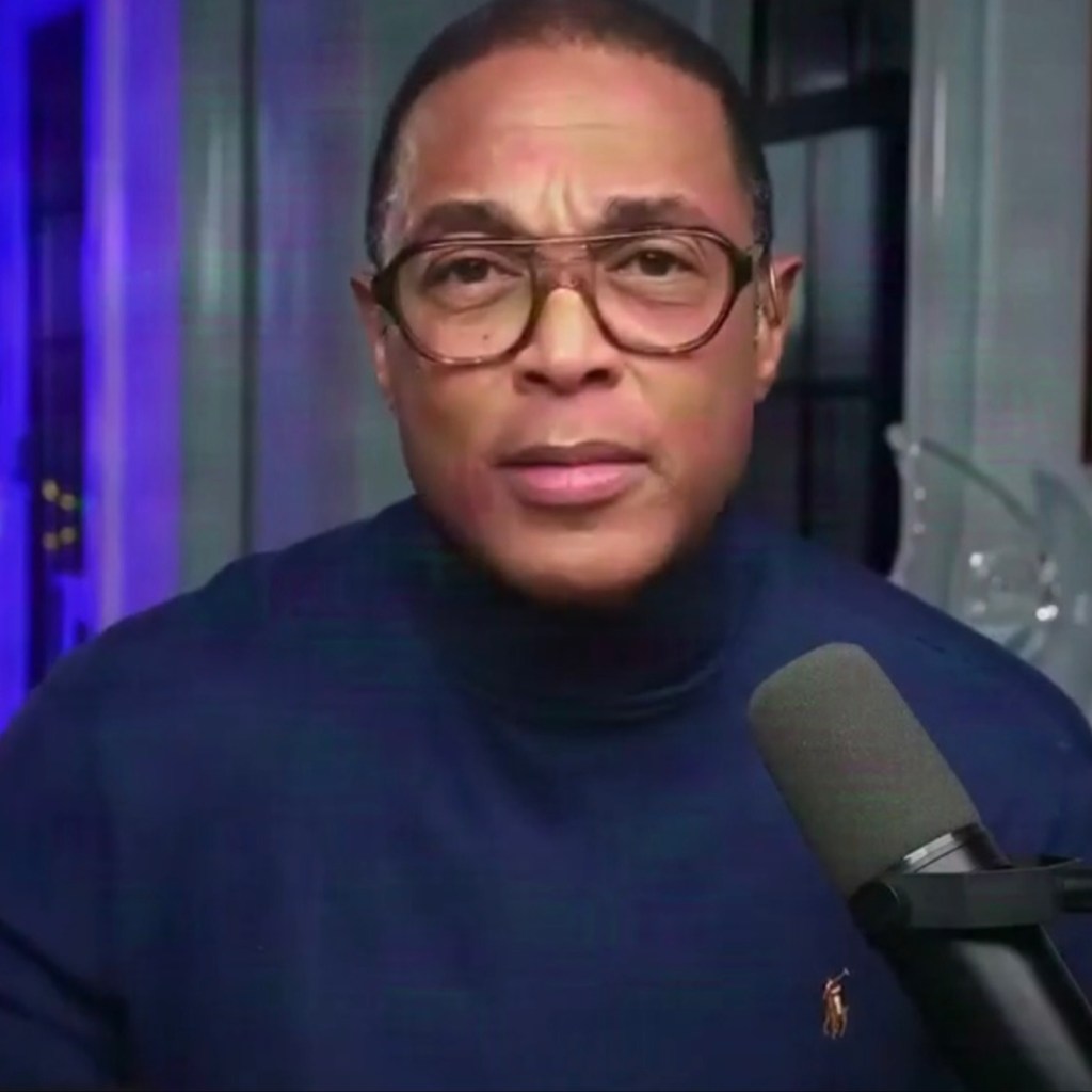Don Lemon wearing glasses and a blue turtleneck while speaking into a microphone.