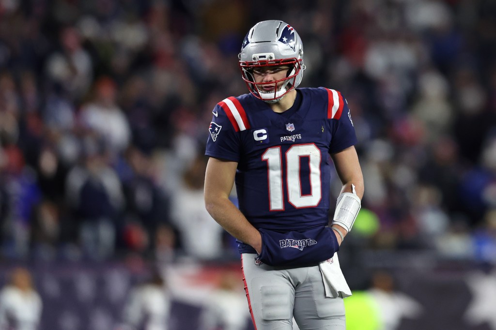 New England Patriots player Mac Jones with jersey number 10, hands in a pouch, wearing a helmet and looking down.