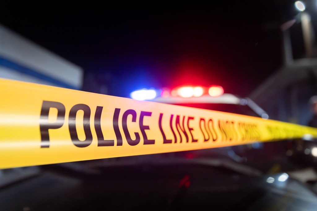 Yellow police tape with "POLICE LINE DO NOT CROSS" written on it, stretched across the foreground with a police car's flashing red and blue lights blurred in the background at night.