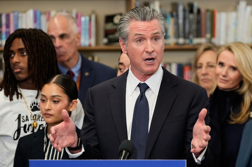California Governor Gavin Newsom speaking at a press conference, gesturing with both hands, with people behind him.