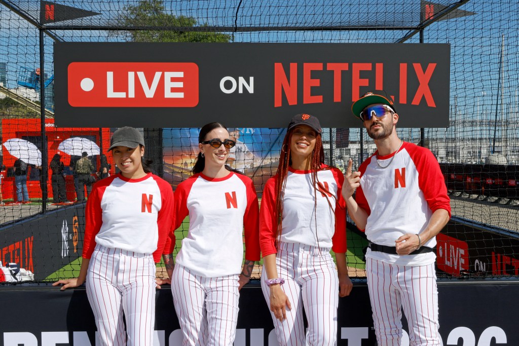 Four people in baseball uniforms with "N" on shirts stand in front of a "LIVE ON NETFLIX" sign at Oracle Park.