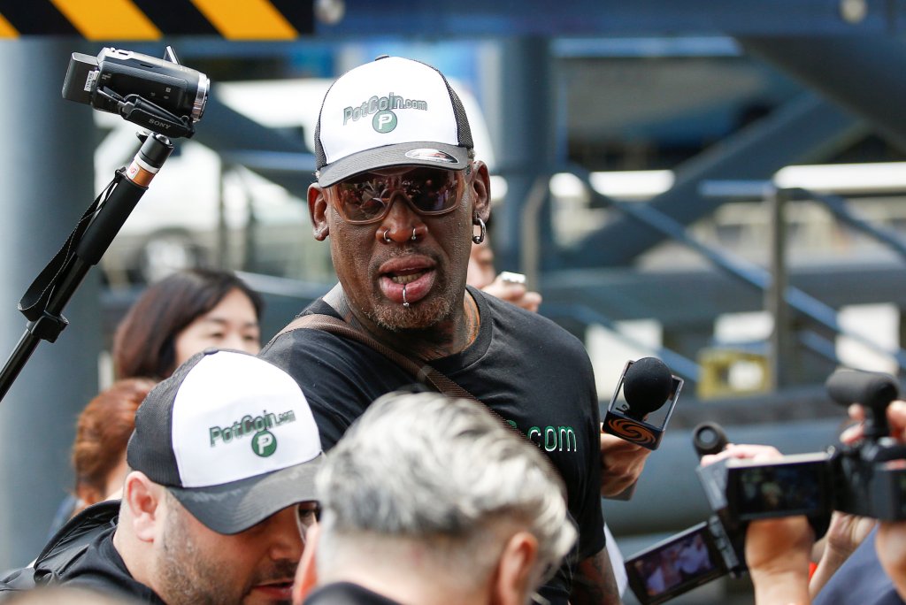 Former NBA player Dennis Rodman, wearing sunglasses and a PotCoin.com hat and shirt, speaks into a microphone at Beijing airport.