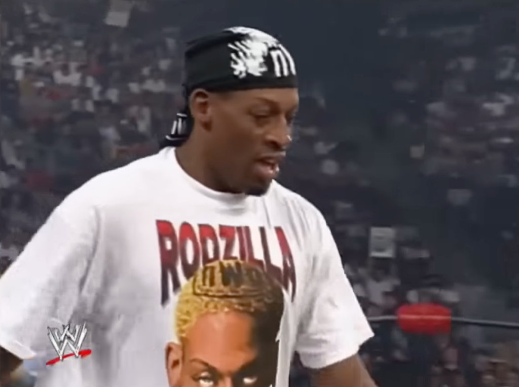 Dennis Rodman in a white "Rodzilla" t-shirt with a blonde-haired graphic, black bandana, in a wrestling ring.