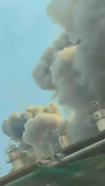 Thick, dark smoke rises into a light blue sky, with white industrial structures visible below.