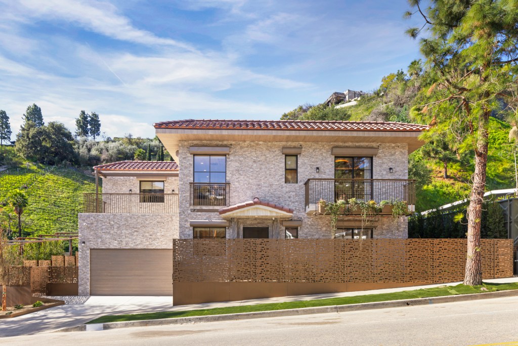 A stone house with a terracotta roof, balconies, and a long patterned metal fence across the front, with a hillside of green trees and bushes behind it.