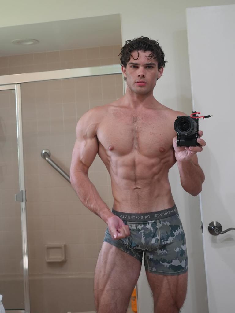 A muscular man in camouflage underwear holding a camera, posing in front of a mirror.