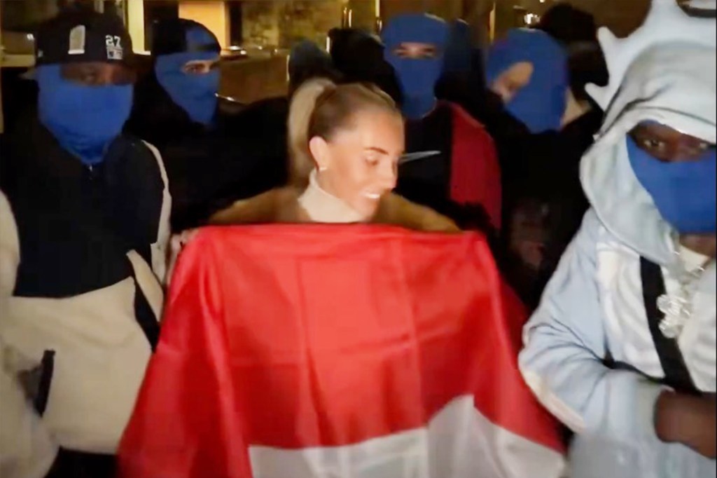 Bonnie Blue (Tia Billinger) smiling in front of the Indonesian Embassy in the UK, surrounded by people in blue balaclavas and holding the Indonesian flag.