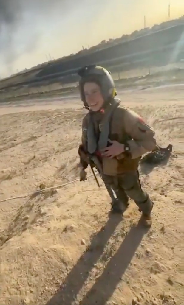 A smiling US pilot in uniform and helmet stands in a sandy, desert-like area, with industrial structures in the background.