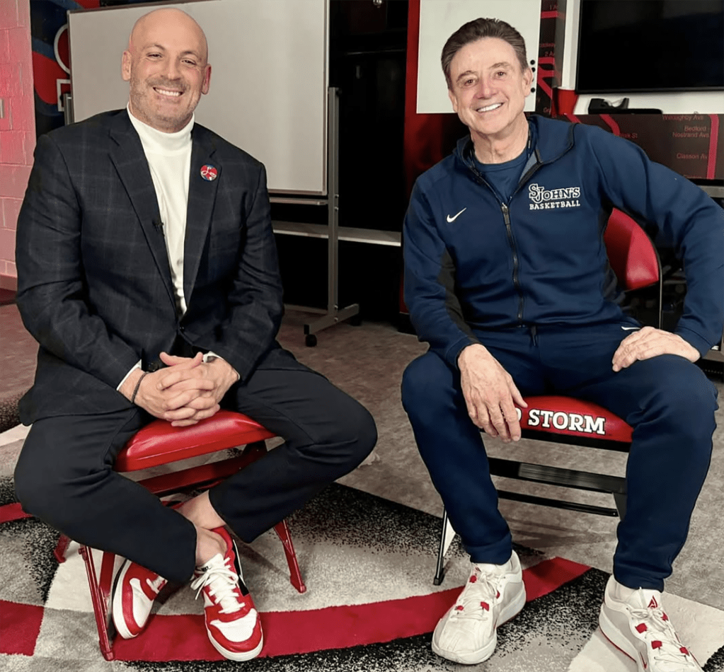 Two men, one in a suit and one in a tracksuit, sit on red chairs.