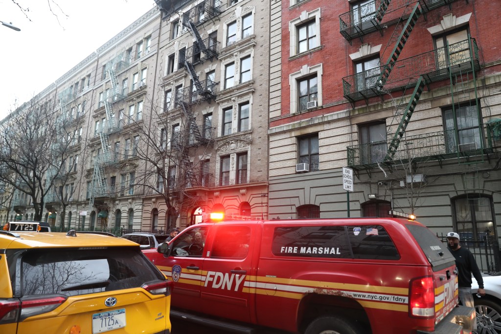 A red FDNY Fire Marshal truck with flashing lights parked in front of apartment buildings with fire escapes.