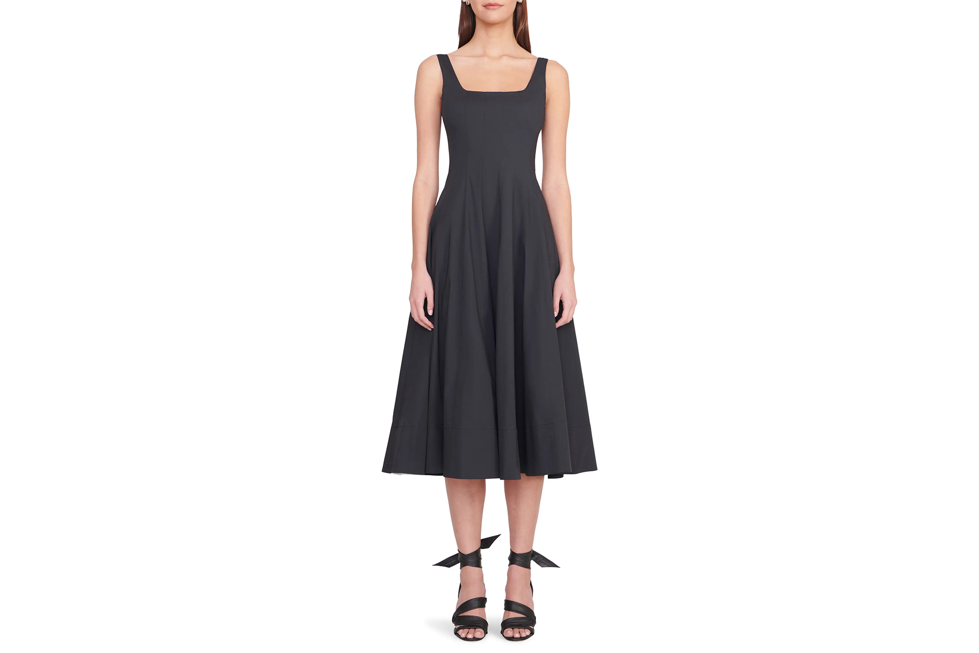 A woman models the Staud Wells Dress in black, a sleeveless midi dress with a square neckline and a full, flared skirt, paired with black ribbon-tie sandals.