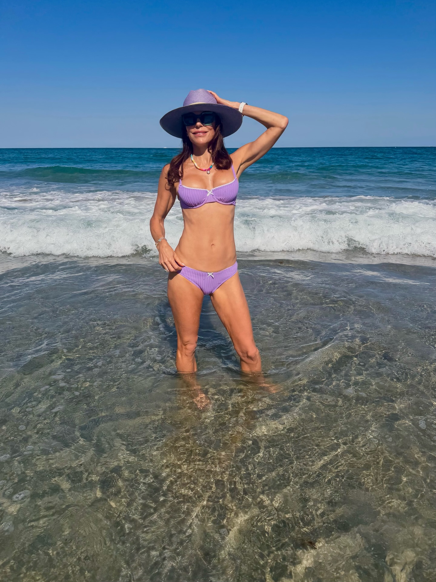 Bethenny Frankel standing in the ocean wearing a purple bikini and sun hat.